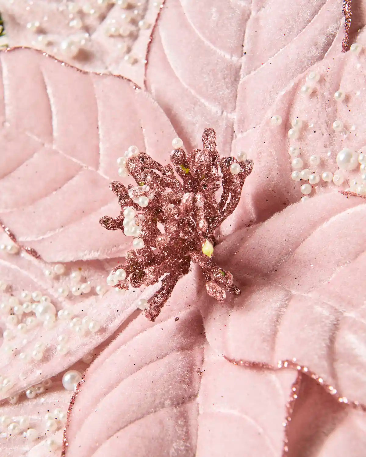 A close-up of a pink fabric surface with quilted leaf-like texture, scattered pearl-like beads, and a glittery rose-gold floral centre.