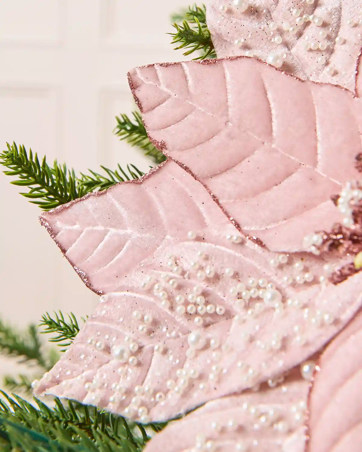 A close-up of a pink poinsettia-style decorative leaf with raised leaf texture and white pearl-like bead accents, shown with green pine branches against a soft light background.