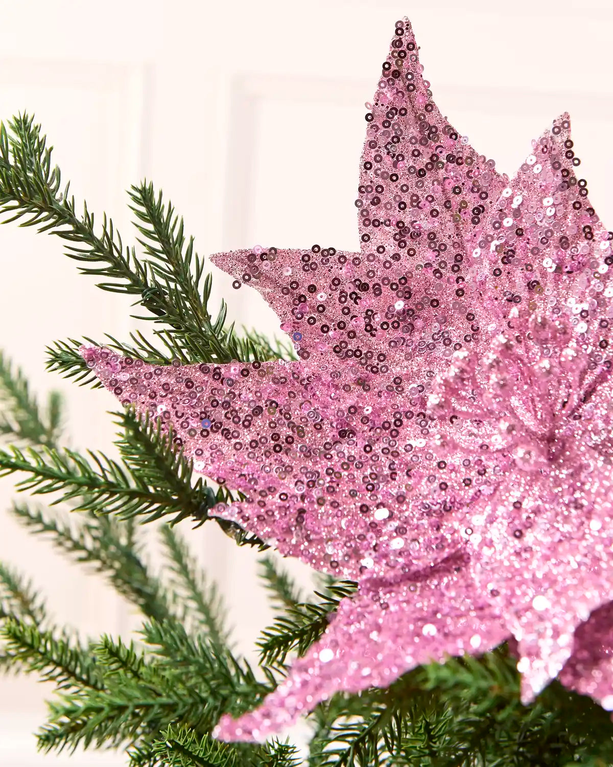 A close-up of a pink sequined star decoration resting on green pine branches against a soft off-white background.