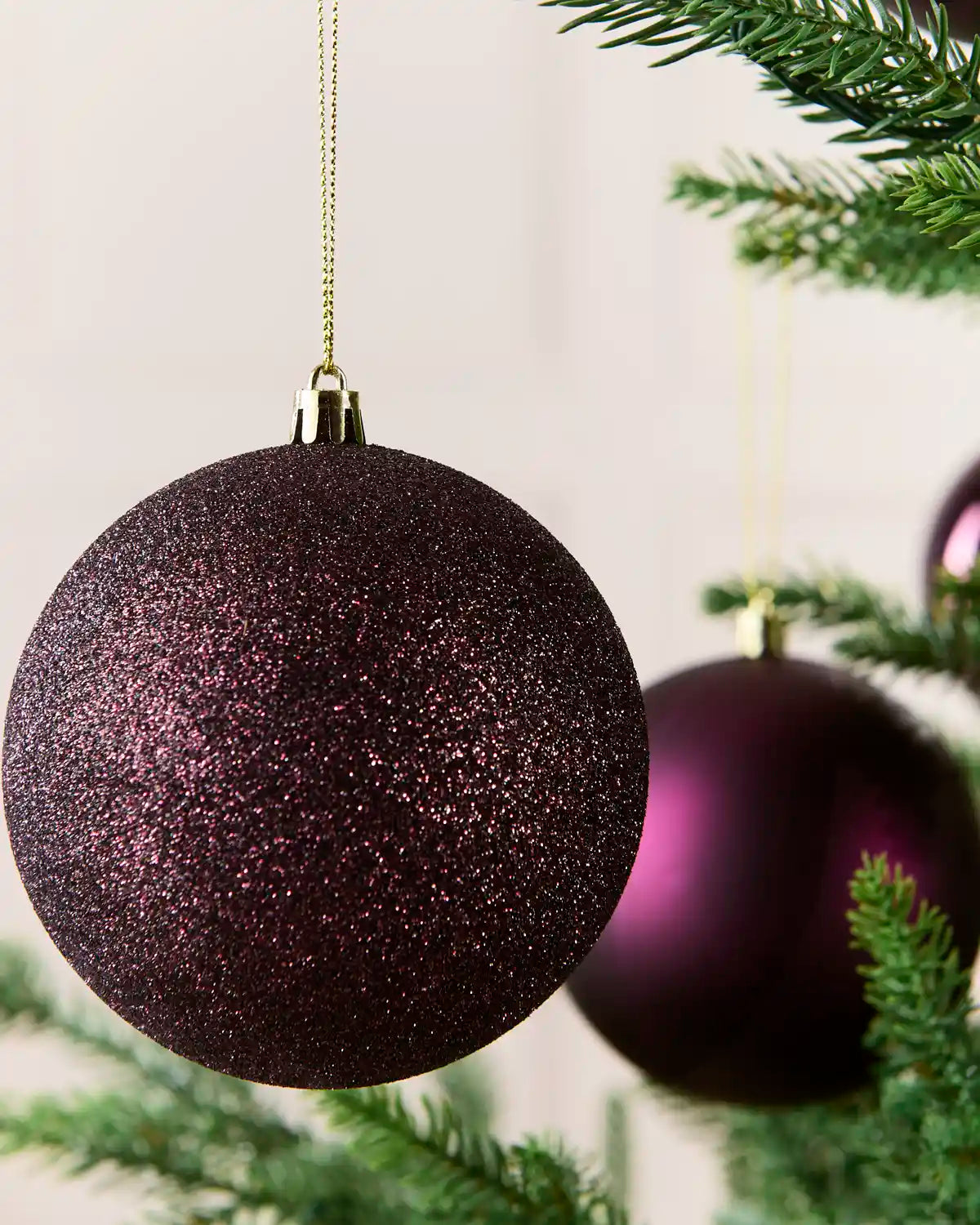 A close-up of a purple glitter Christmas bauble hanging from a gold string on a Christmas tree, with another purple ornament blurred in the background.