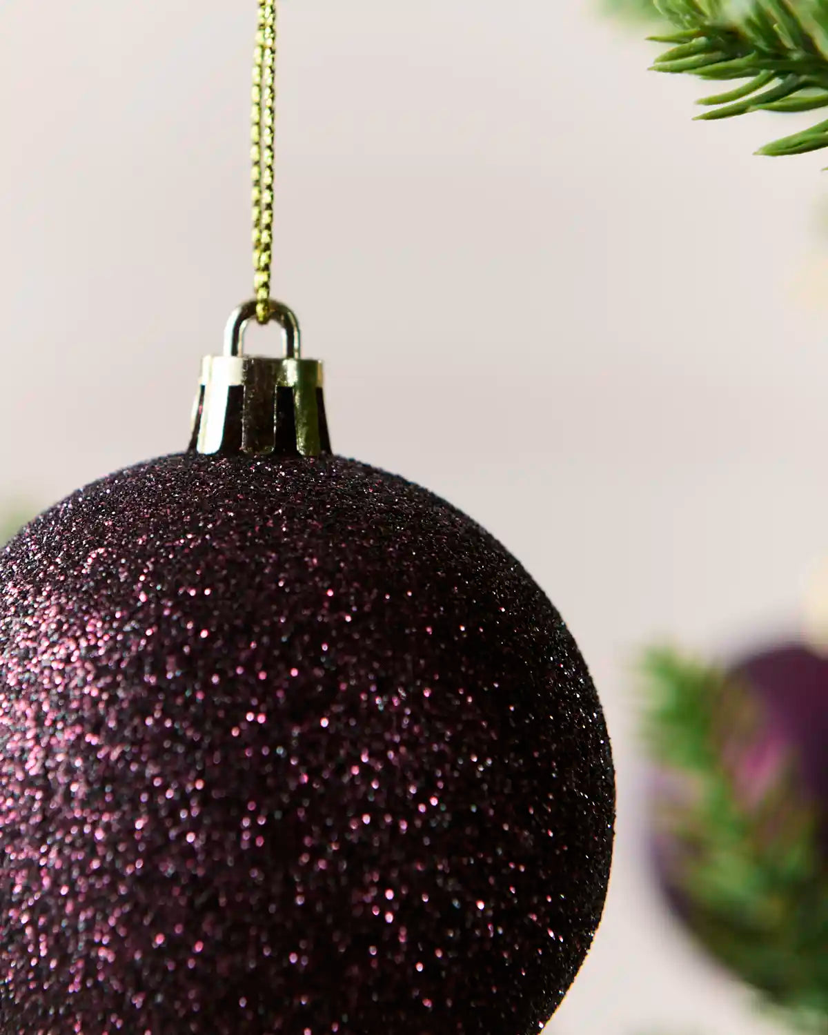 A close-up of a purple glitter Christmas ornament hanging from a gold chain, with blurred pine needles in the background.