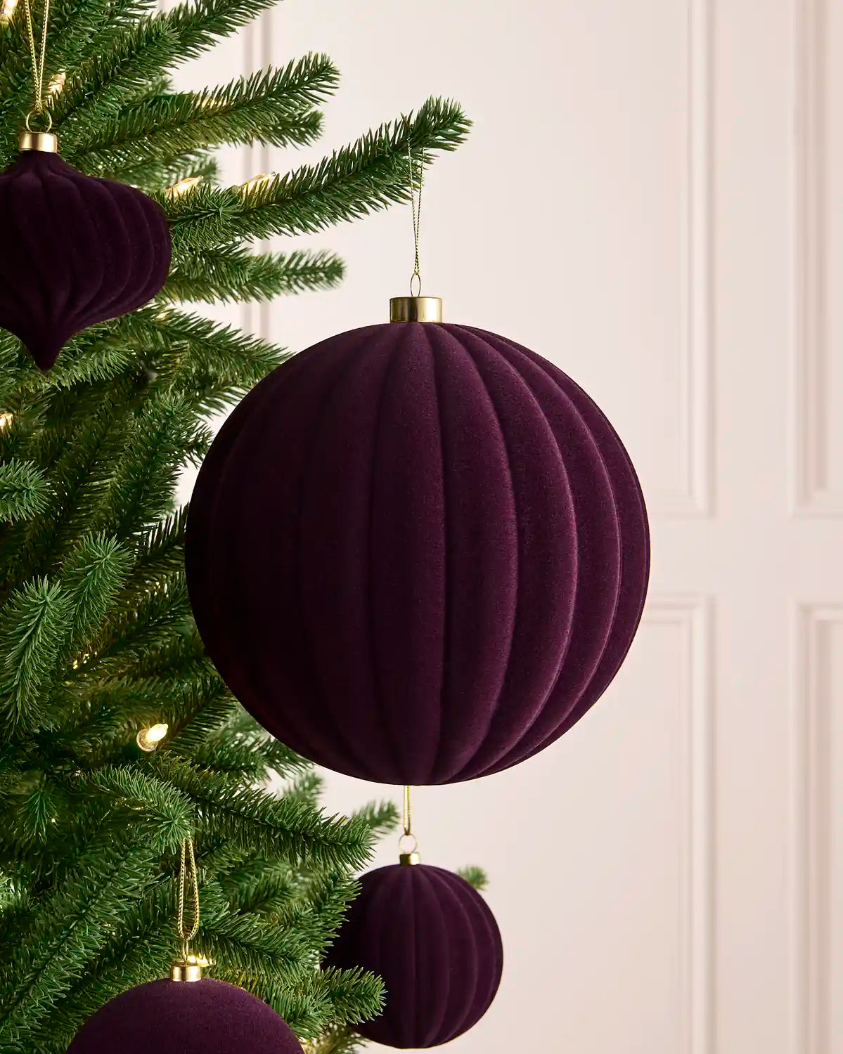 A close-up of a purple ribbed velvet Christmas tree ornament hanging from a green pine tree branch, with other matching ornaments and warm string lights in the background.
