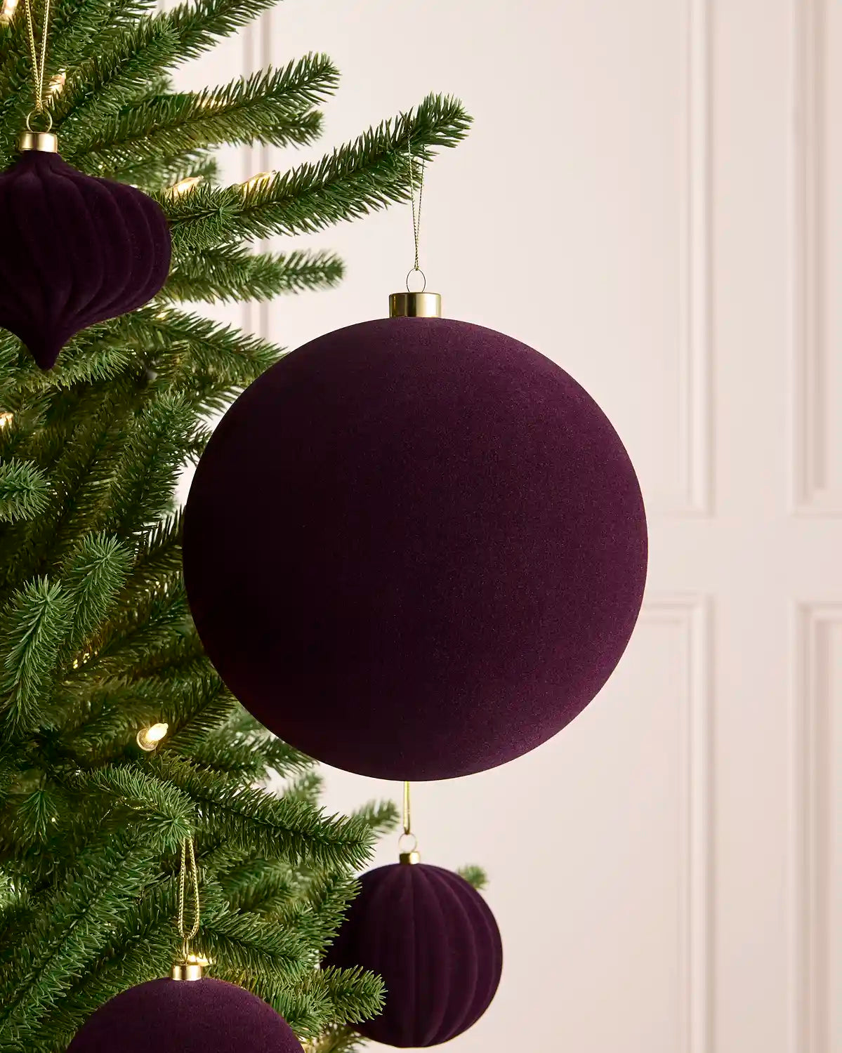 A close-up of a purple velvet Christmas tree ornament hanging on a green artificial Christmas tree, with a soft cream panelled wall in the background.