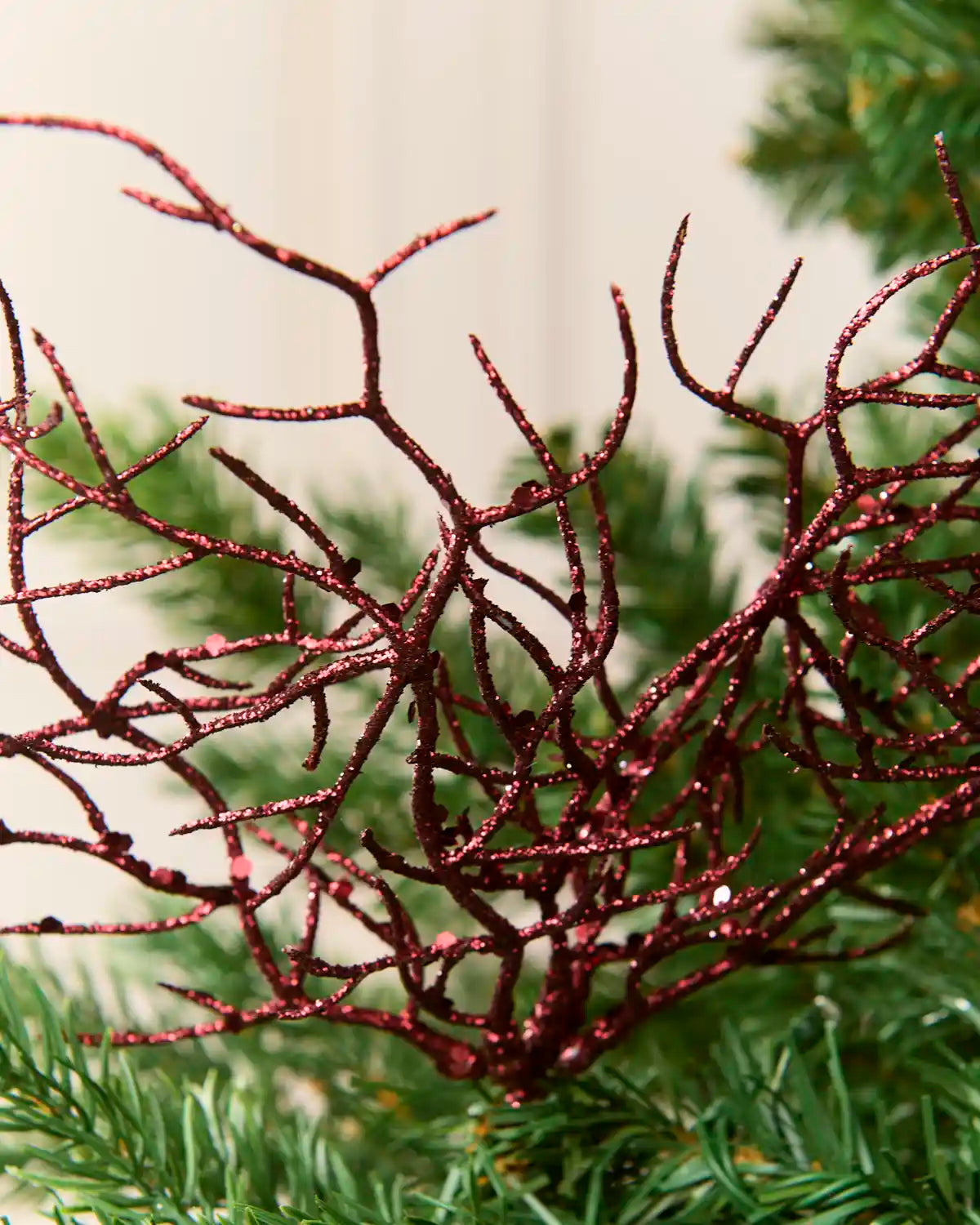 A close-up of a red glittery coral branch decorative topper resting on green pine needles against a soft beige background.