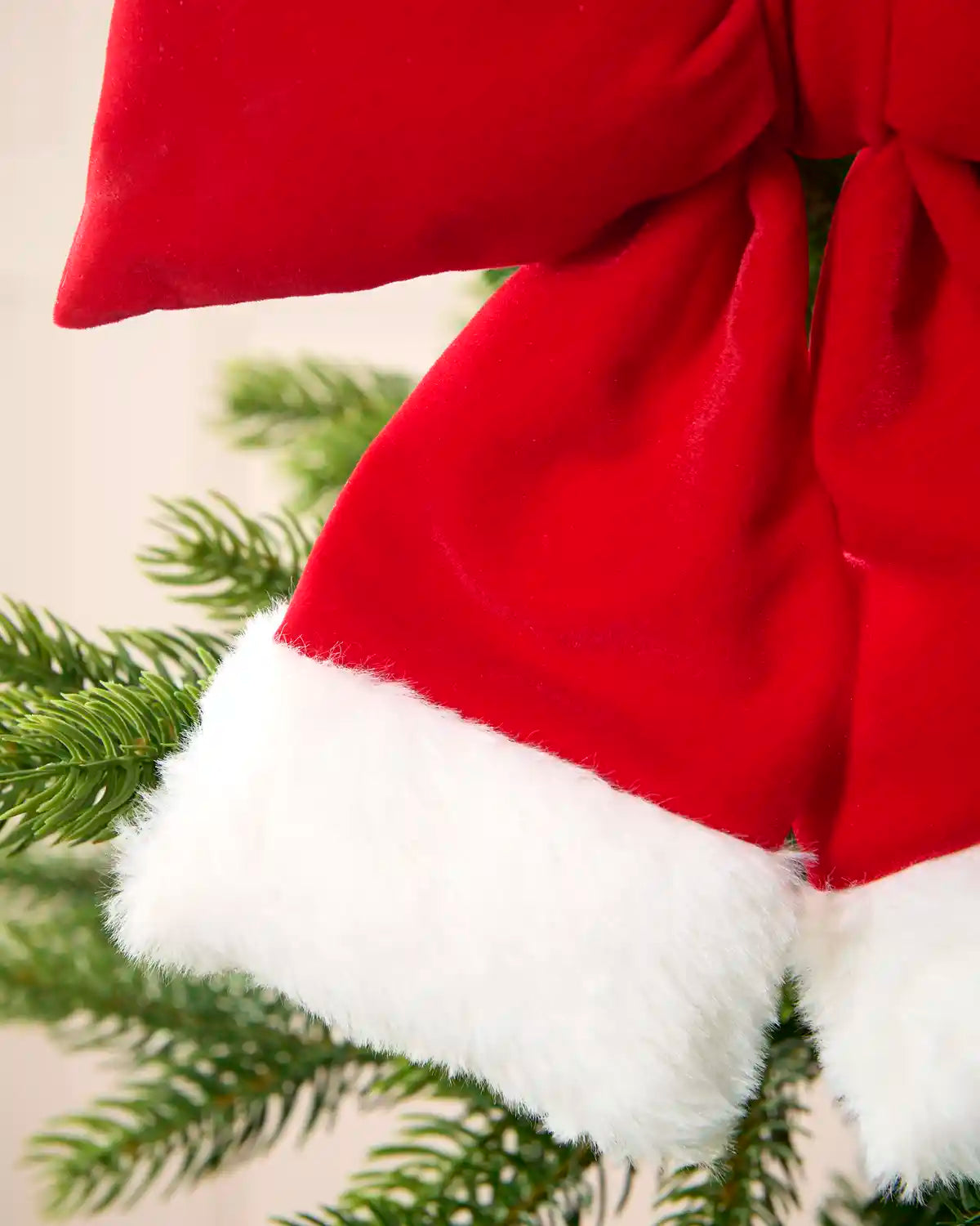 A close-up of a red velvet Christmas tree ornament with a white faux fur trim, styled on a green pine tree branch.