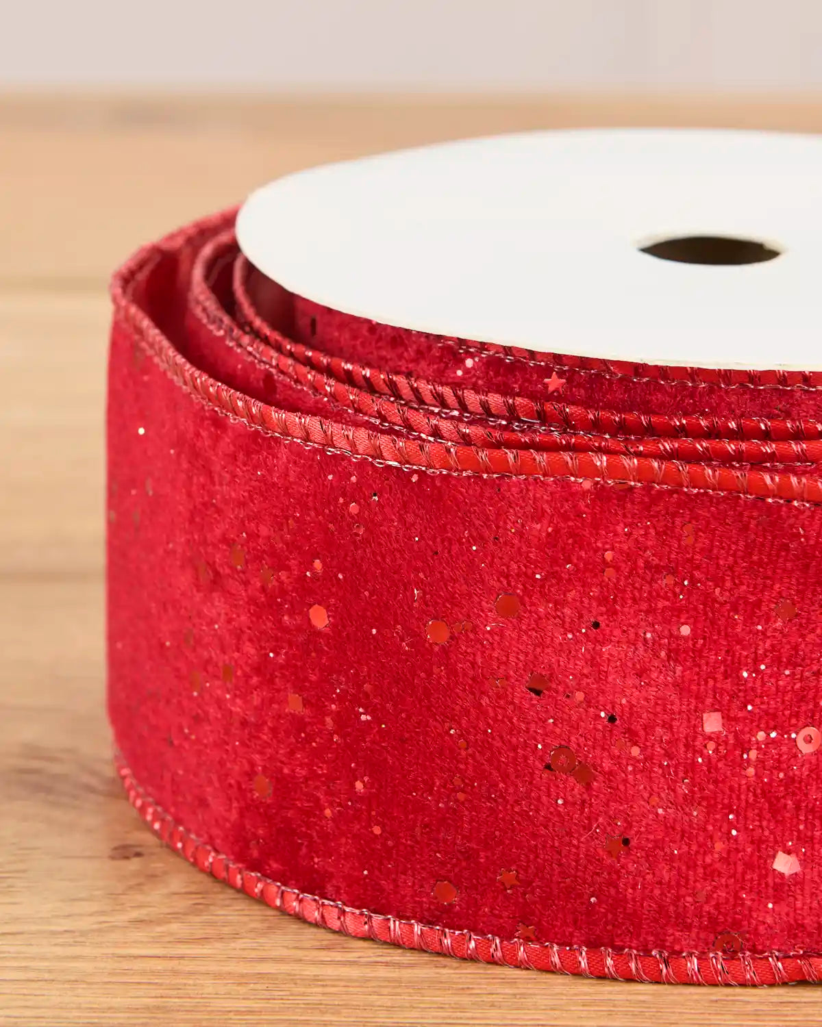 A close-up of a red velvet ribbon with small red glitter specks, wound on a white spool over a light wood surface.