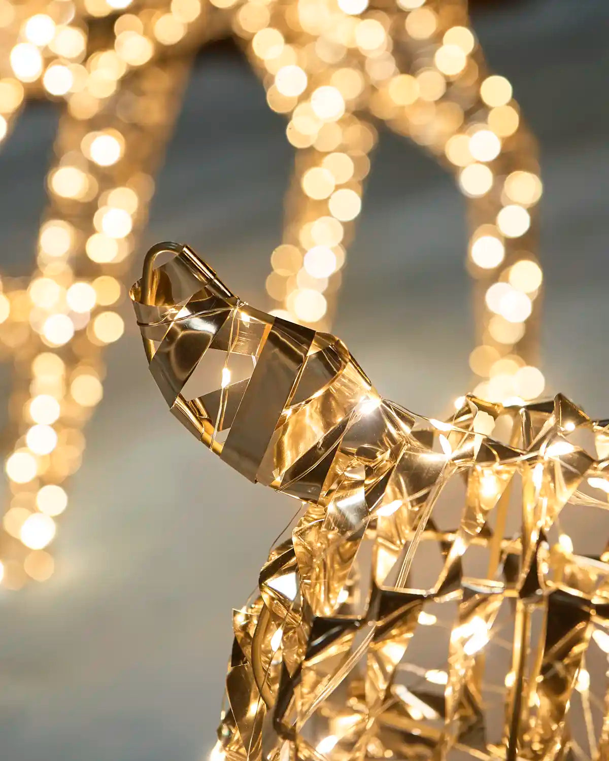 A close-up of a reindeers LED tail, geometric golden deer sculpture illuminated with warm lights, set against a soft-focus background of glowing bokeh holiday string lights.