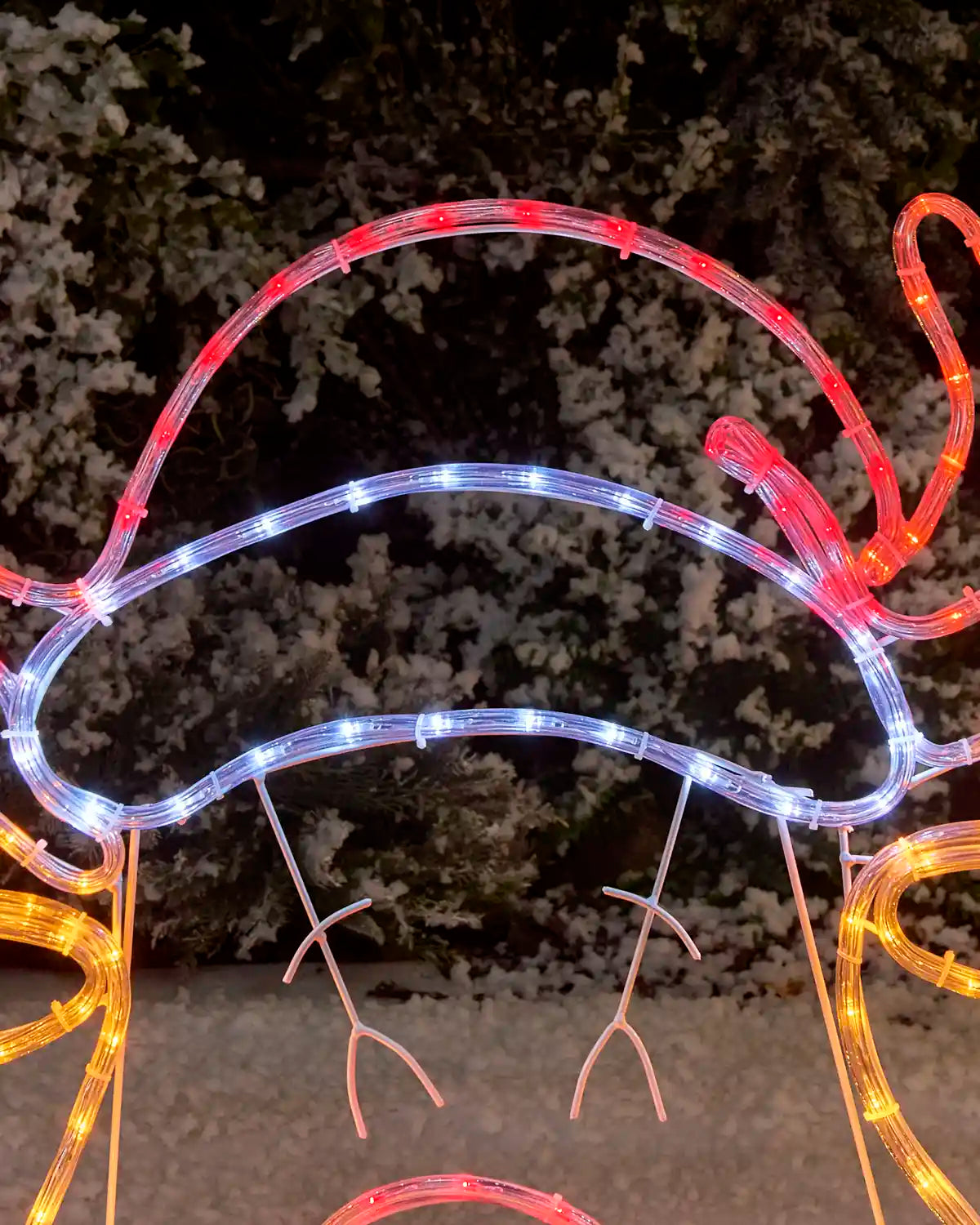 A close-up of a reindeers Santa hat LED light decoration glowing in red, set against a dark, blurred outdoor background.