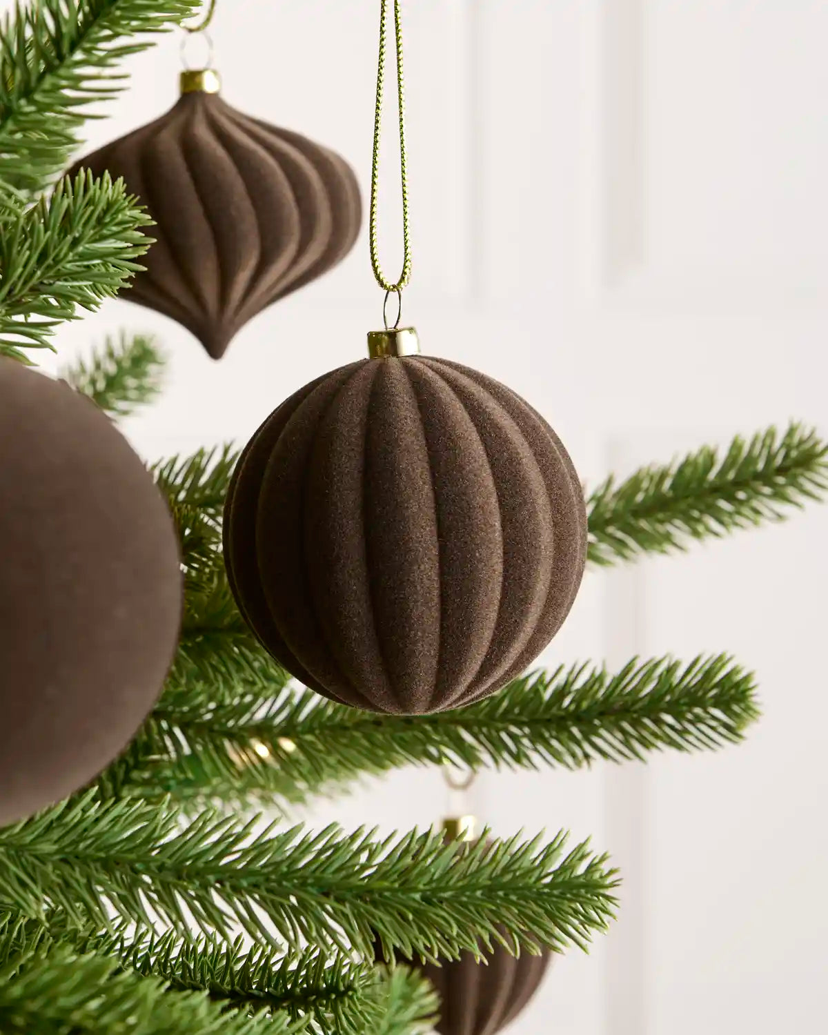A close-up of a ribbed brown felt Christmas ornament hanging from a green pine tree branch against a soft white background.