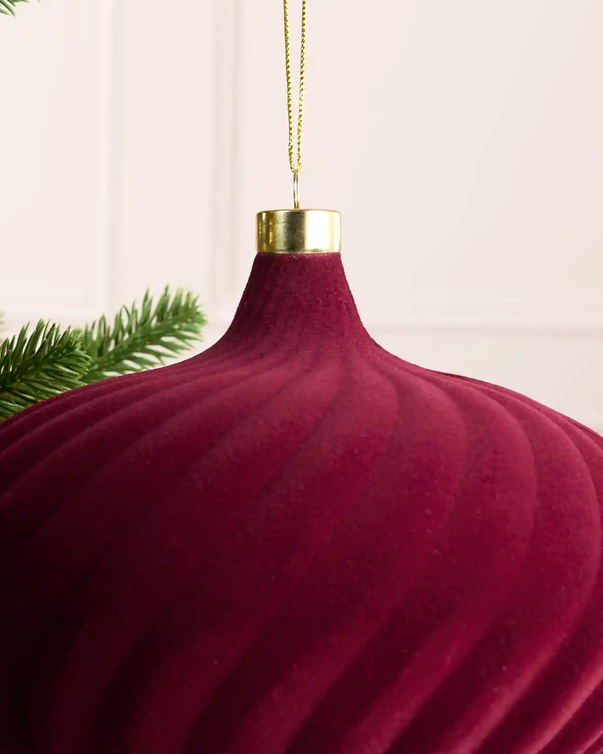 A close-up of a ribbed, burgundy Christmas ornament with a gold cap and hanging loop, shown against a soft cream background with a hint of green pine branches.