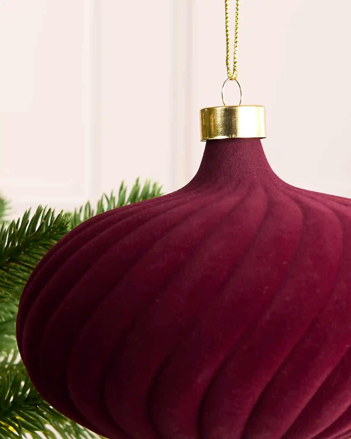 A close-up of a ribbed, deep burgundy Christmas ornament with a gold cap and gold hanging string, partially framed by green pine branches against a soft off-white background.