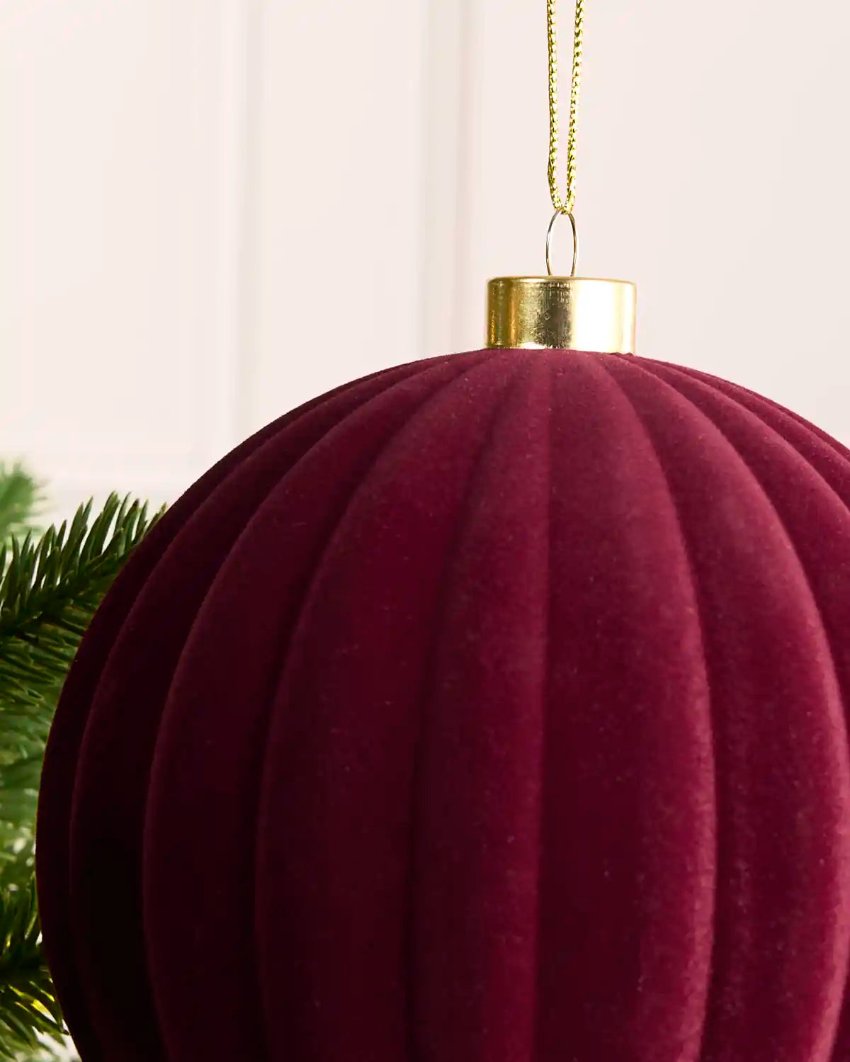 A close-up of a ribbed, pleated dark burgundy velvet Christmas ornament with a gold metal cap and hanging loop, shown next to green pine needles against a soft off-white background.