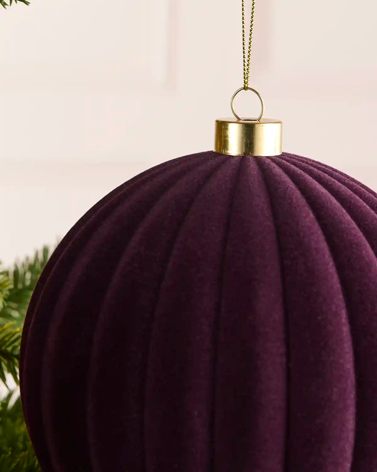 A close-up of a ribbed, purple velvet Christmas ornament hanging from a gold cap with a gold chain against a soft light background.