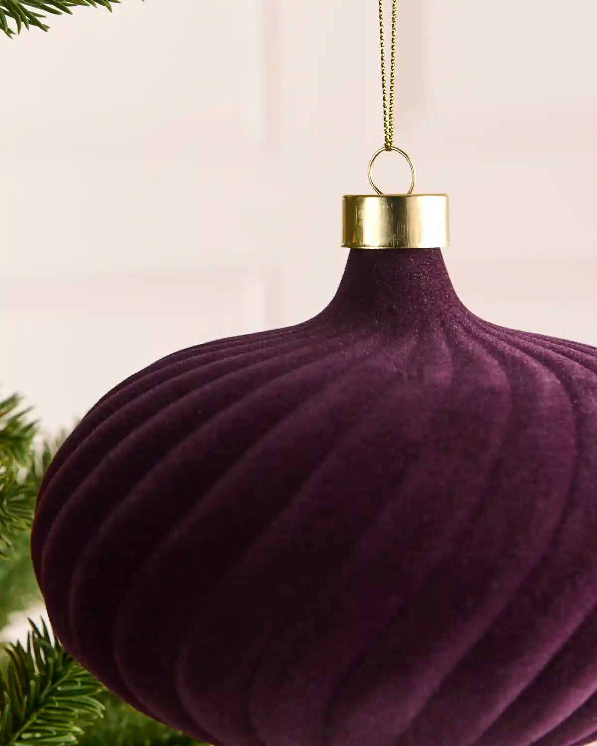 A close-up of a ribbed, purple velvet Christmas ornament with a gold cap and hanging loop, shown hanging near green pine branches against a soft cream background.