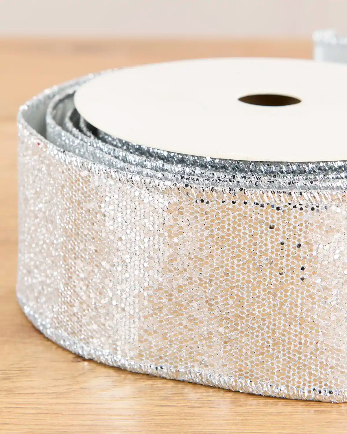 A close-up of a roll of glittery silver sequin ribbon on a light wood surface, showing the reflective texture and pattern of the ribbon.