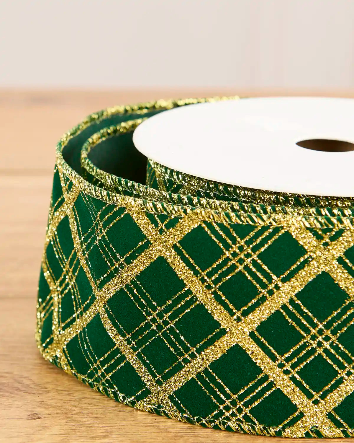 A close-up of a roll of green ribbon with a glittery gold plaid pattern, wound around a white spool on a light wooden surface.