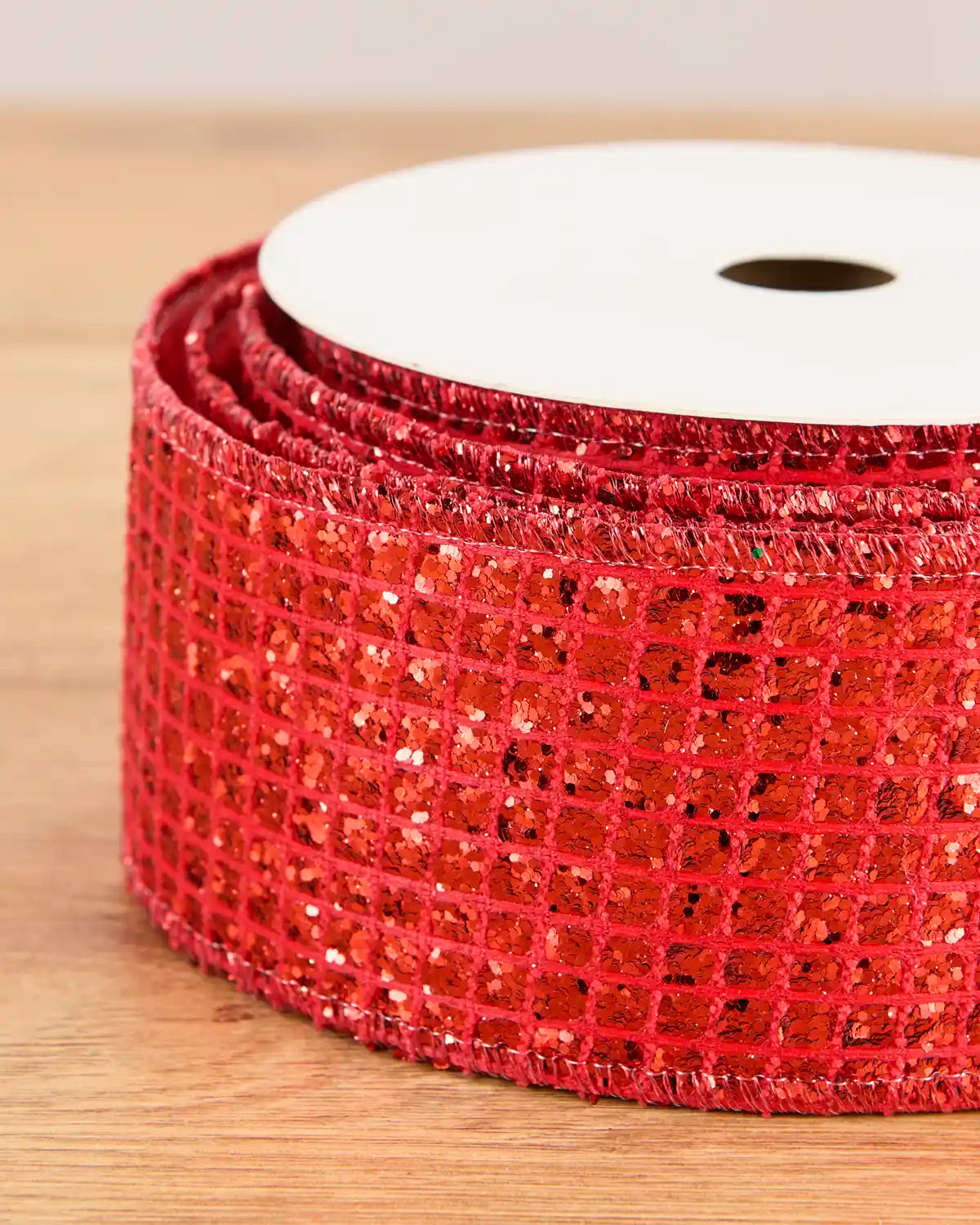 A close-up of a roll of red sequin mesh ribbon on a white spool, set on a light wooden surface.