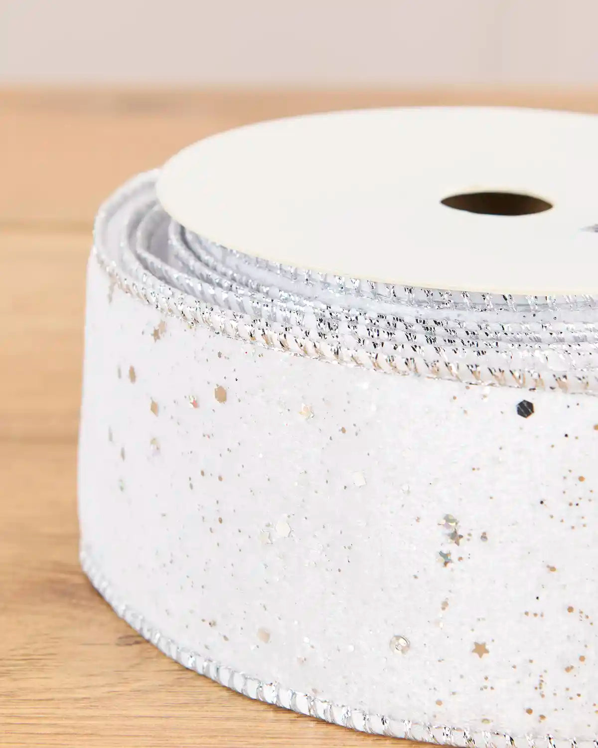 A close-up of a roll of white glitter ribbon with silver and gold star-shaped sequins, wound on a white spool and resting on a light wooden surface.
