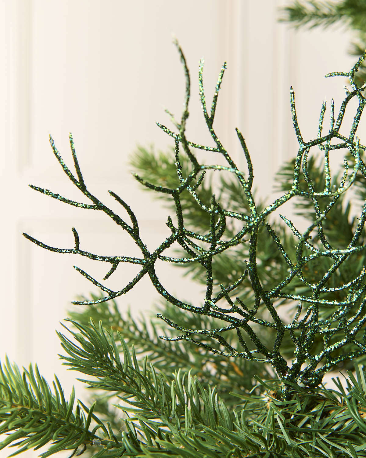 A close-up of a sage green glittery decorative branch or artificial pine accent layered over real green pine needles, against a soft cream paneled background.