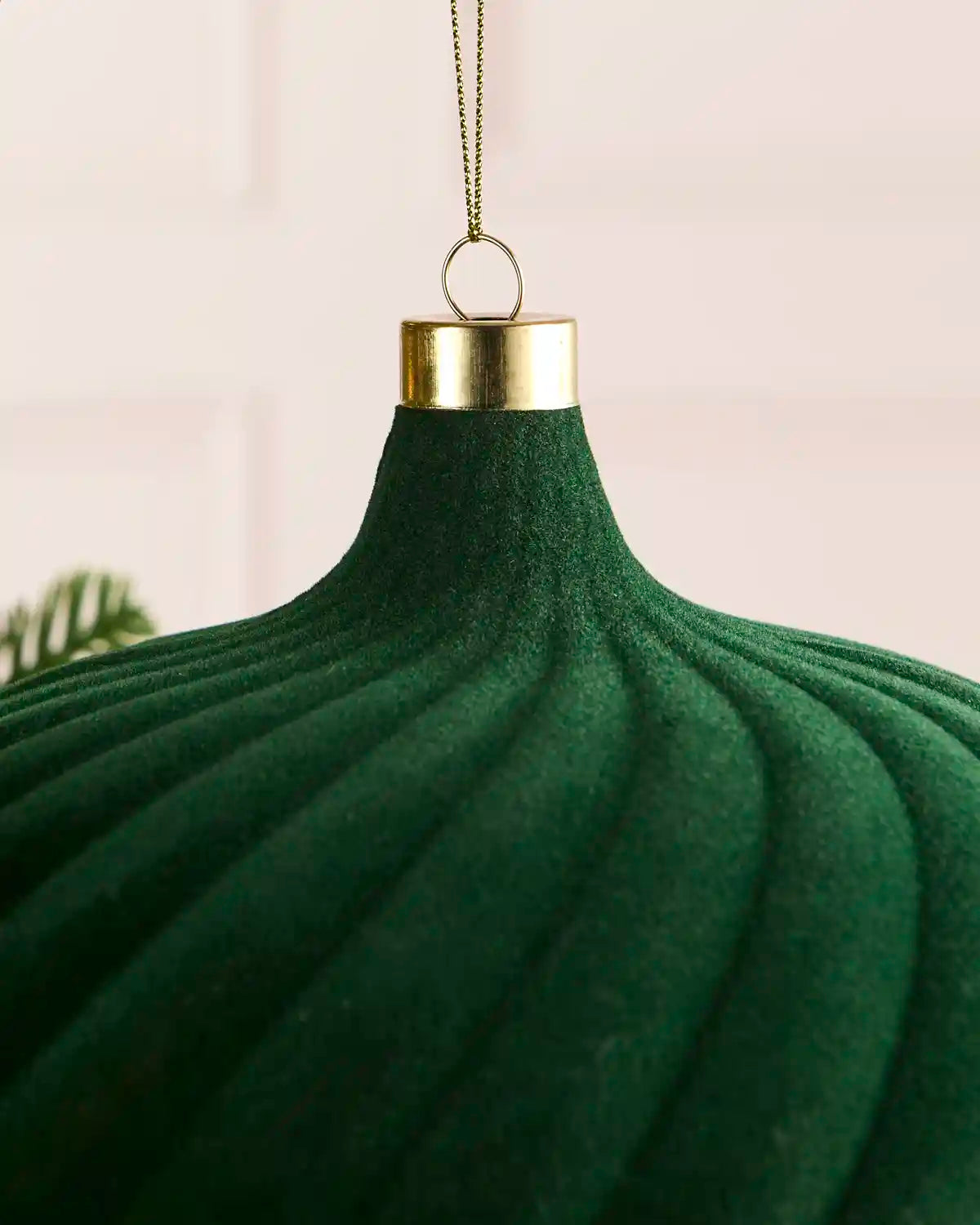 A close-up of a sage green ribbed Christmas ornament with a gold metal cap and hanging loop, shown against a soft light pink background.
