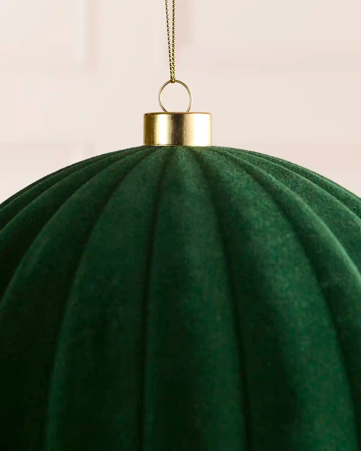 A close-up of a sage green ribbed fabric ornament or hanging decoration with a metallic gold cap and loop, suspended by a thin gold chain against a soft light background.