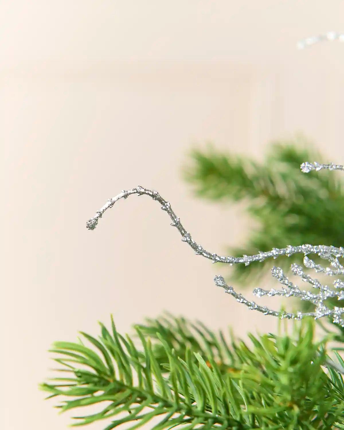 A close-up of a silver glittery decorative branch or ornament resting on green pine needles against a soft beige background.