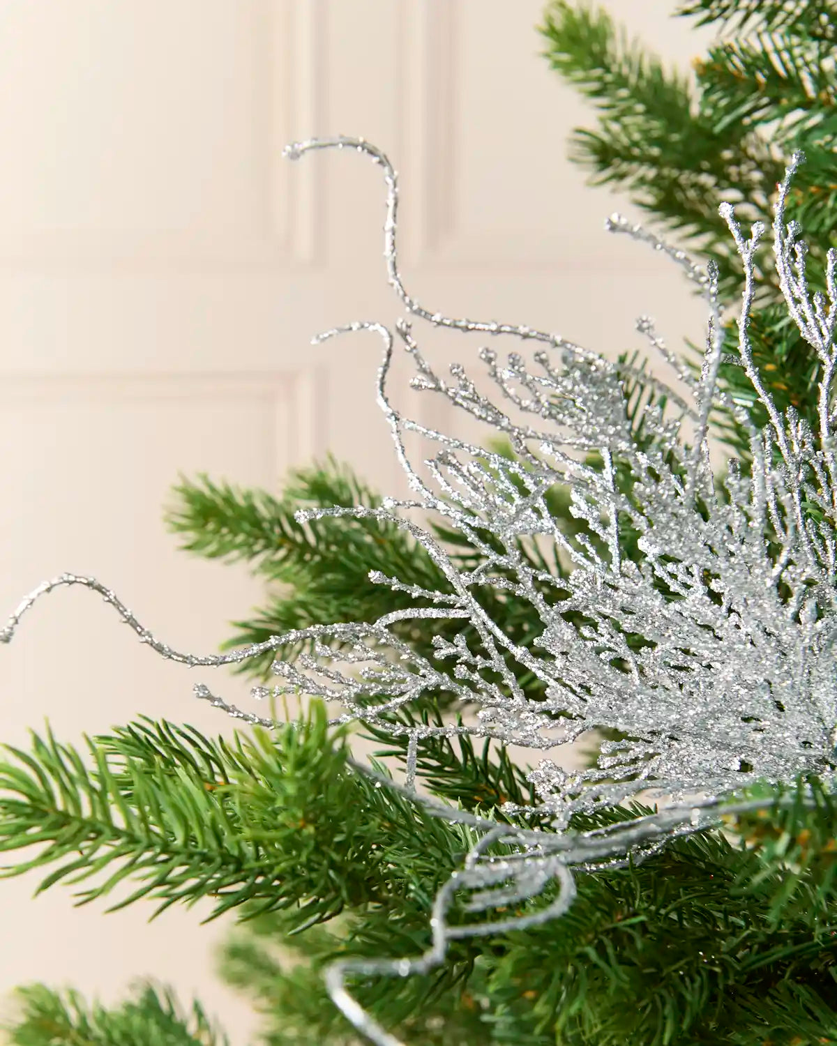 A close-up of a silver glittery decorative branch or pine ornament draped over green evergreen tree branches against a soft beige paneled background.