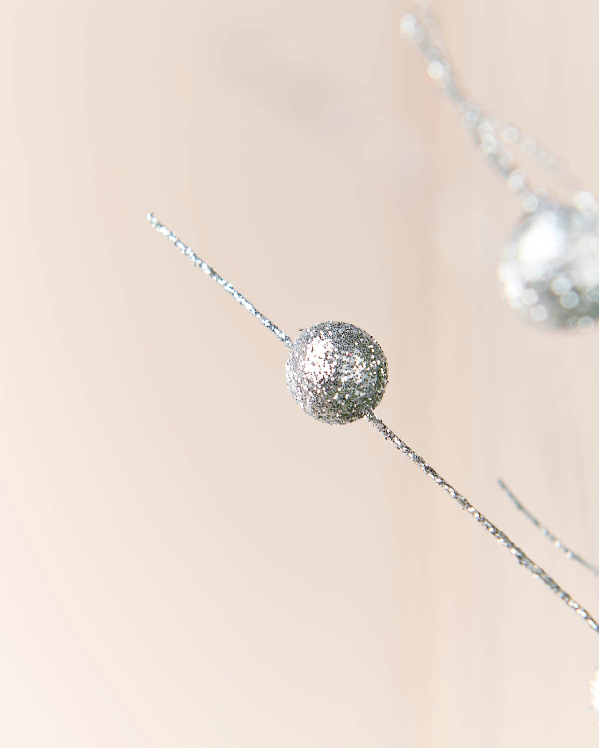 A close-up of a silver glittery spherical ornament hanging from a thin silver wire against a soft beige background.