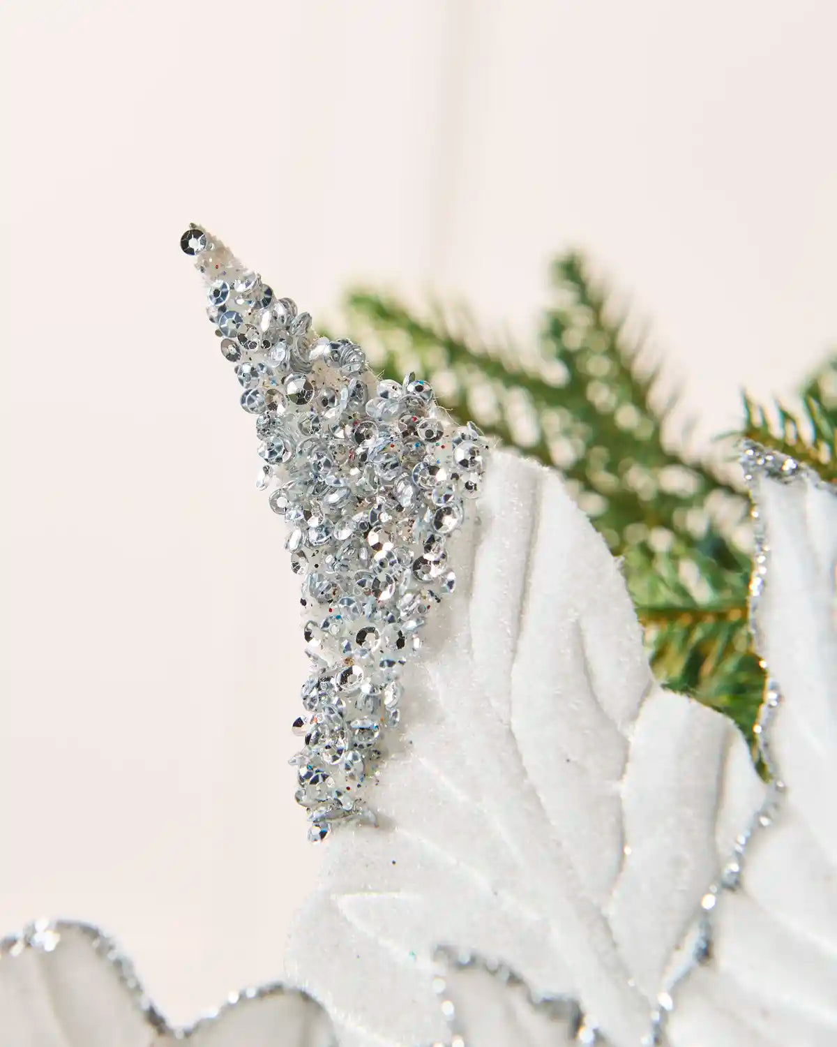 A close-up of a silver rhinestone-embellished Christmas tree poinsettia 
resting on a white textured surface with green pine branches in the background.