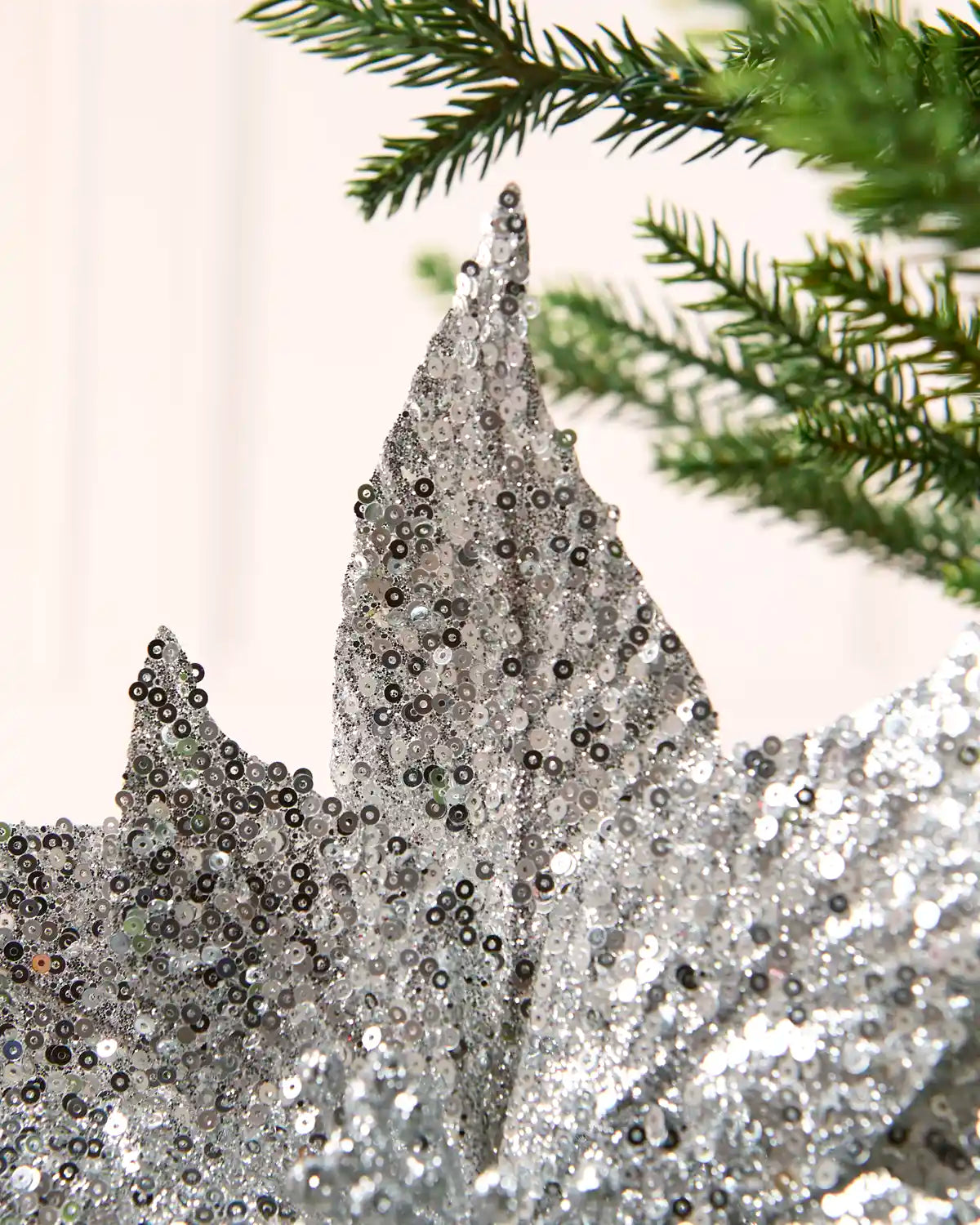 A close-up of a silver sequined decorative poinsettia with green pine branches in the background.