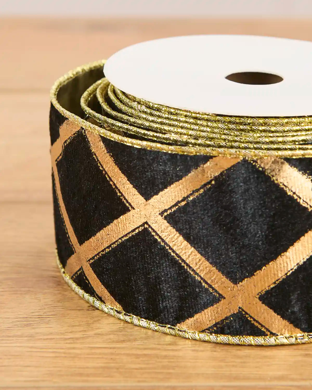 A close-up of a spool of black grosgrain ribbon with a metallic gold crisscross lattice pattern and a twisted gold edge, resting on a light wood surface.