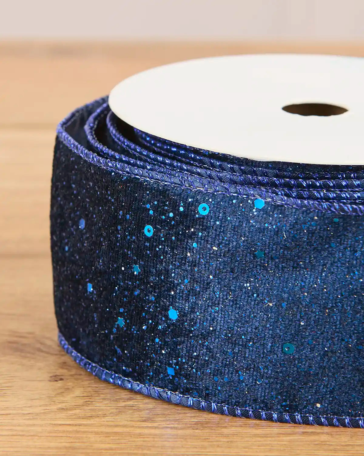 A close-up of a spool of dark blue glitter ribbon with bright blue sequins, resting on a light wooden surface.