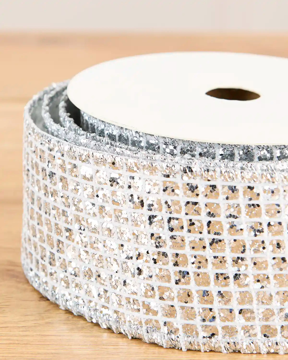 A close-up of a spool of decorative mesh ribbon with silver glitter and small black speckles on a white mesh base, resting on a light wooden surface.