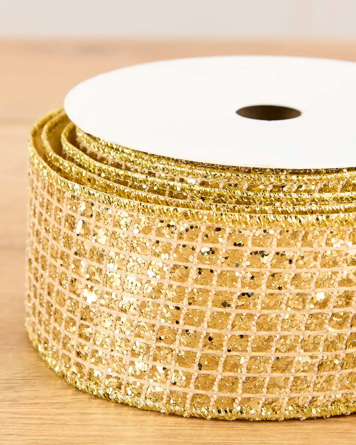 A close-up of a spool of gold glitter mesh ribbon on a light wood surface.