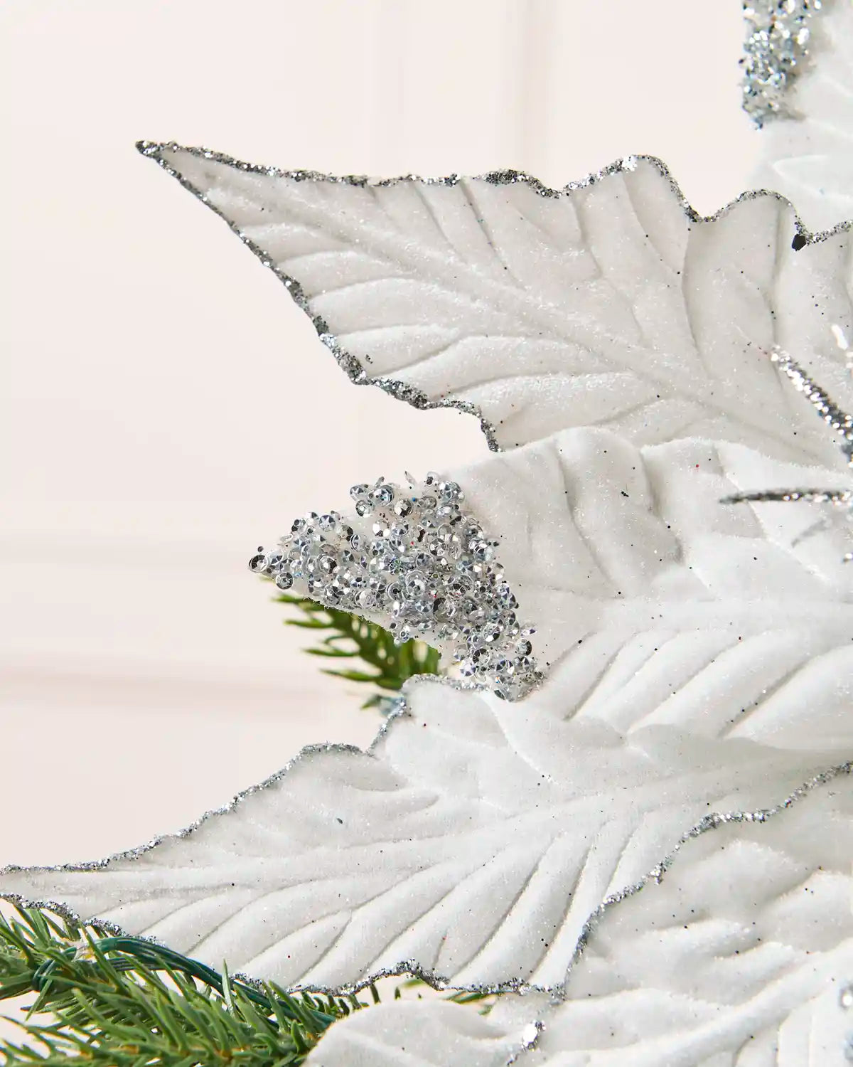 A close-up of a white decorative  poinsettia leaf with silver glitter edging and a sparkling silver cluster accent, shown against a soft light background with green pine branches at the bottom.