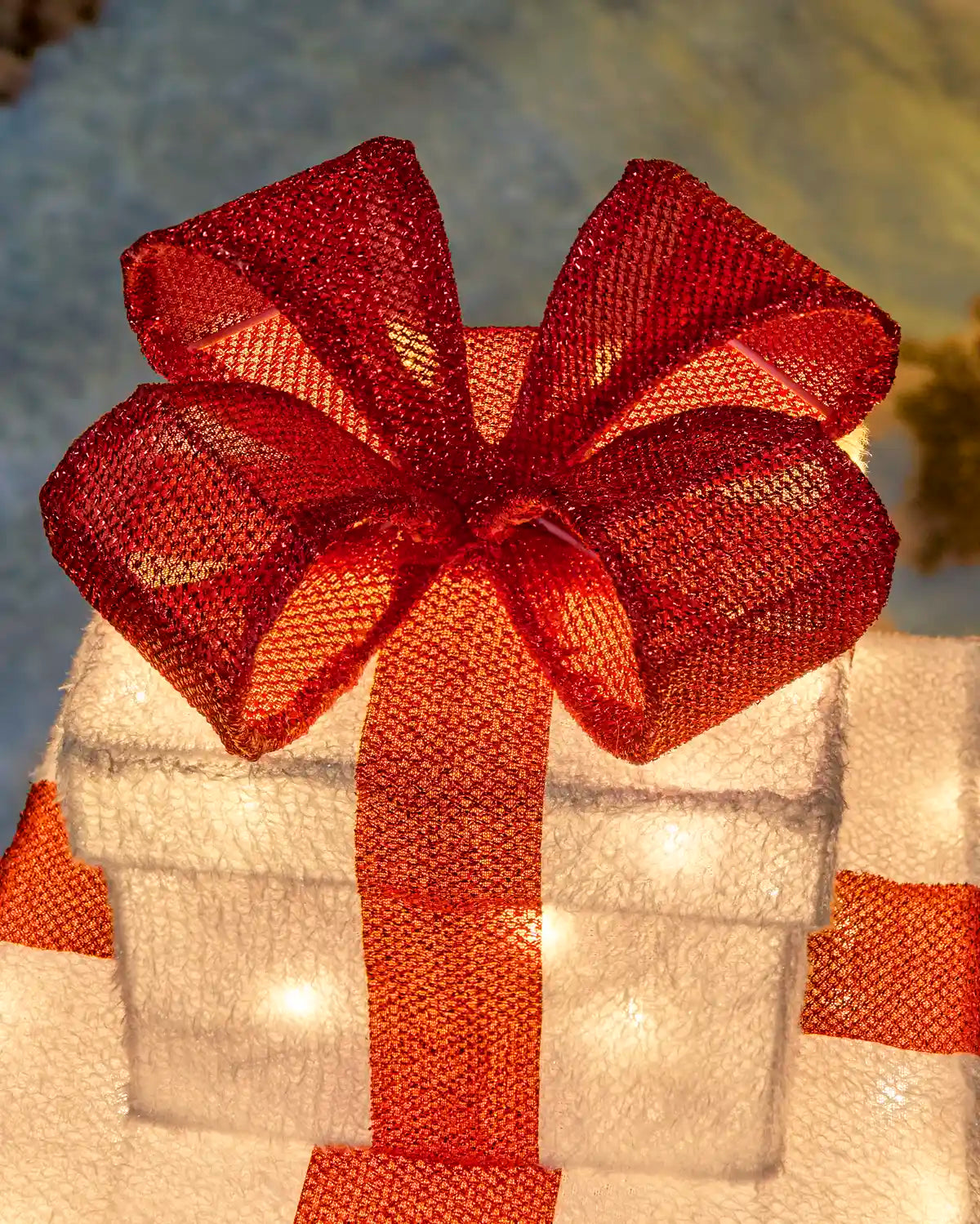 A close-up of a white gift box decorated with a large red mesh bow and red ribbon, with warm white string lights glowing inside, set against a softly blurred outdoor background.