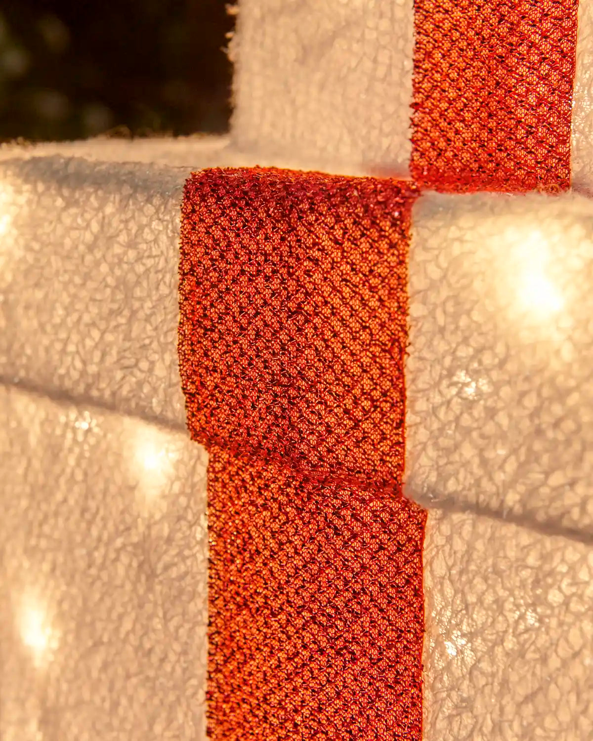 A close-up of a white textured foam Christmas wreath decorated with a wide, shimmering red glittery ribbon, illuminated by warm string lights.