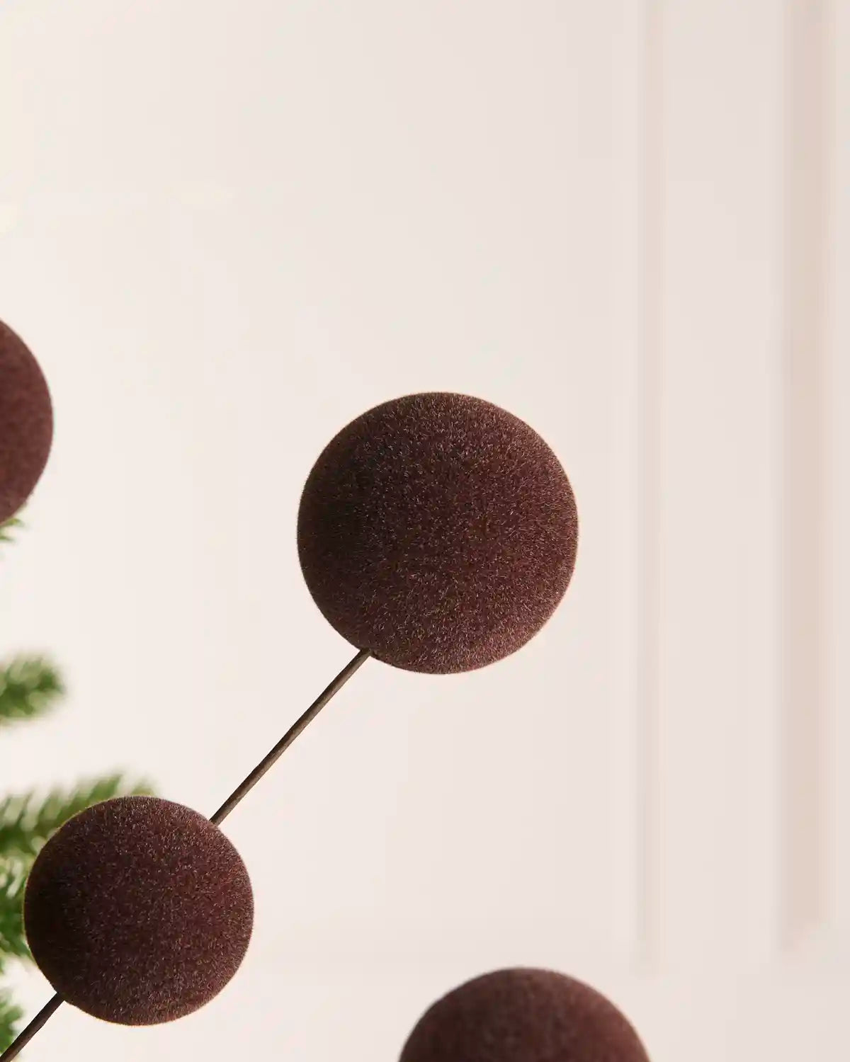 A close-up of an espresso coloured velvet bauble pick ornaments on thin black stems, shown against a soft cream background with a hint of green foliage on the left.
