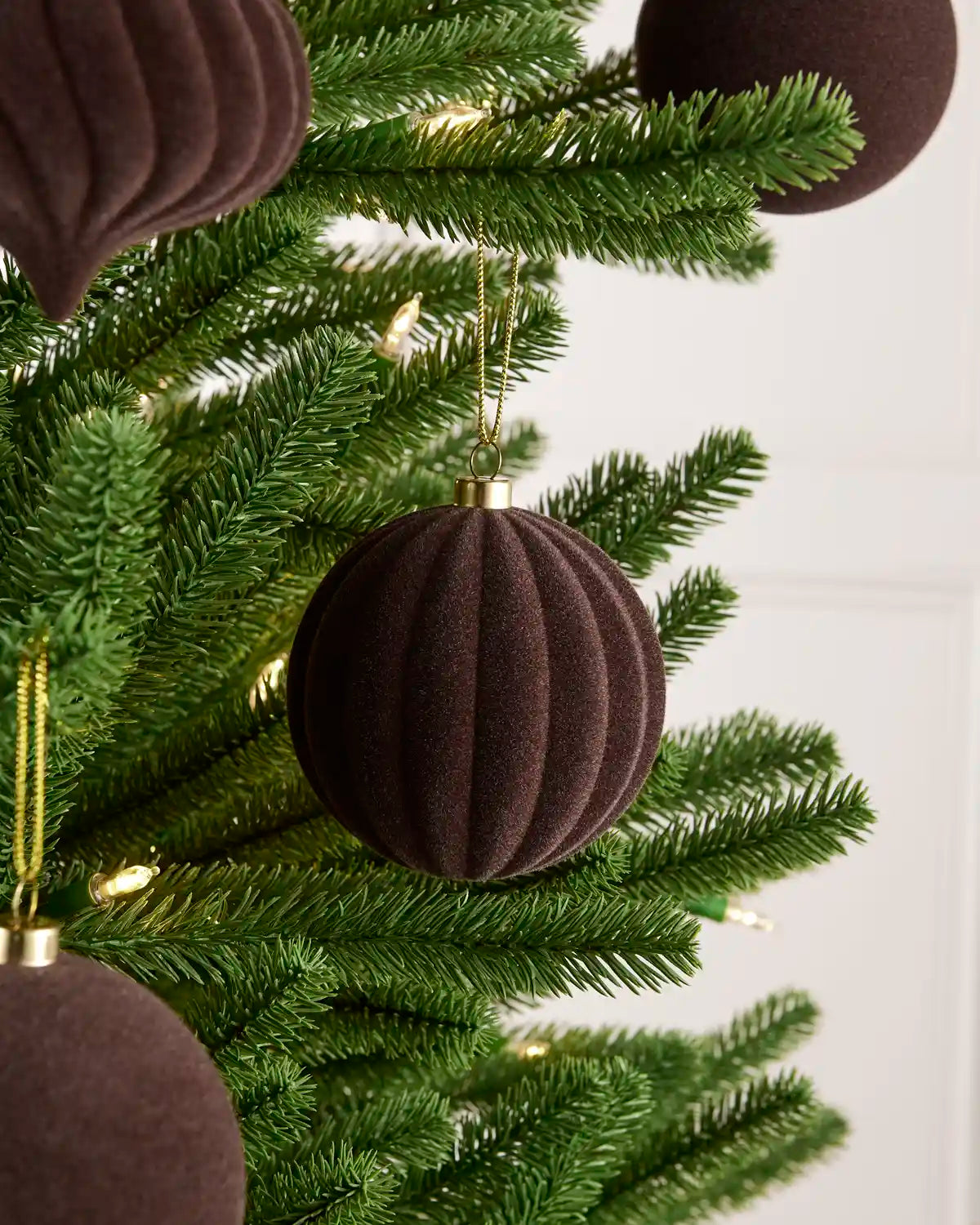 A close-up of an espresso ribbed velvet Christmas ornament hanging on a green artificial Christmas tree with warm white string lights.