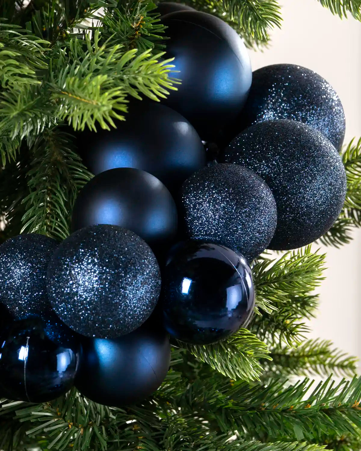 A close-up of dark blue Christmas tree ornaments in three finishes—matte, glitter, and glossy—hanging on a green pine tree branch.
