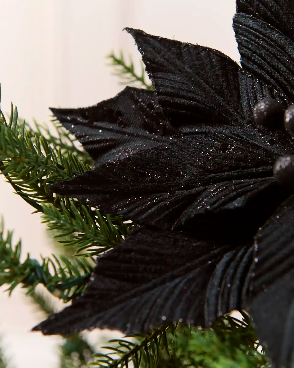 A close-up of the side of a black artificial poinsettia flower with glittery texture, shown next to green pine branches against a light neutral background.