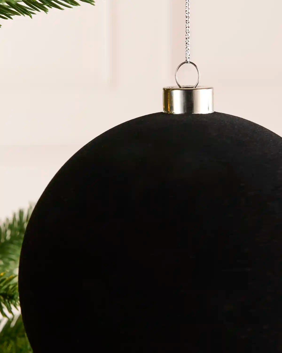 A close up velvet black Christmas bauble hanging from a green Christmas tree branch with a light background.