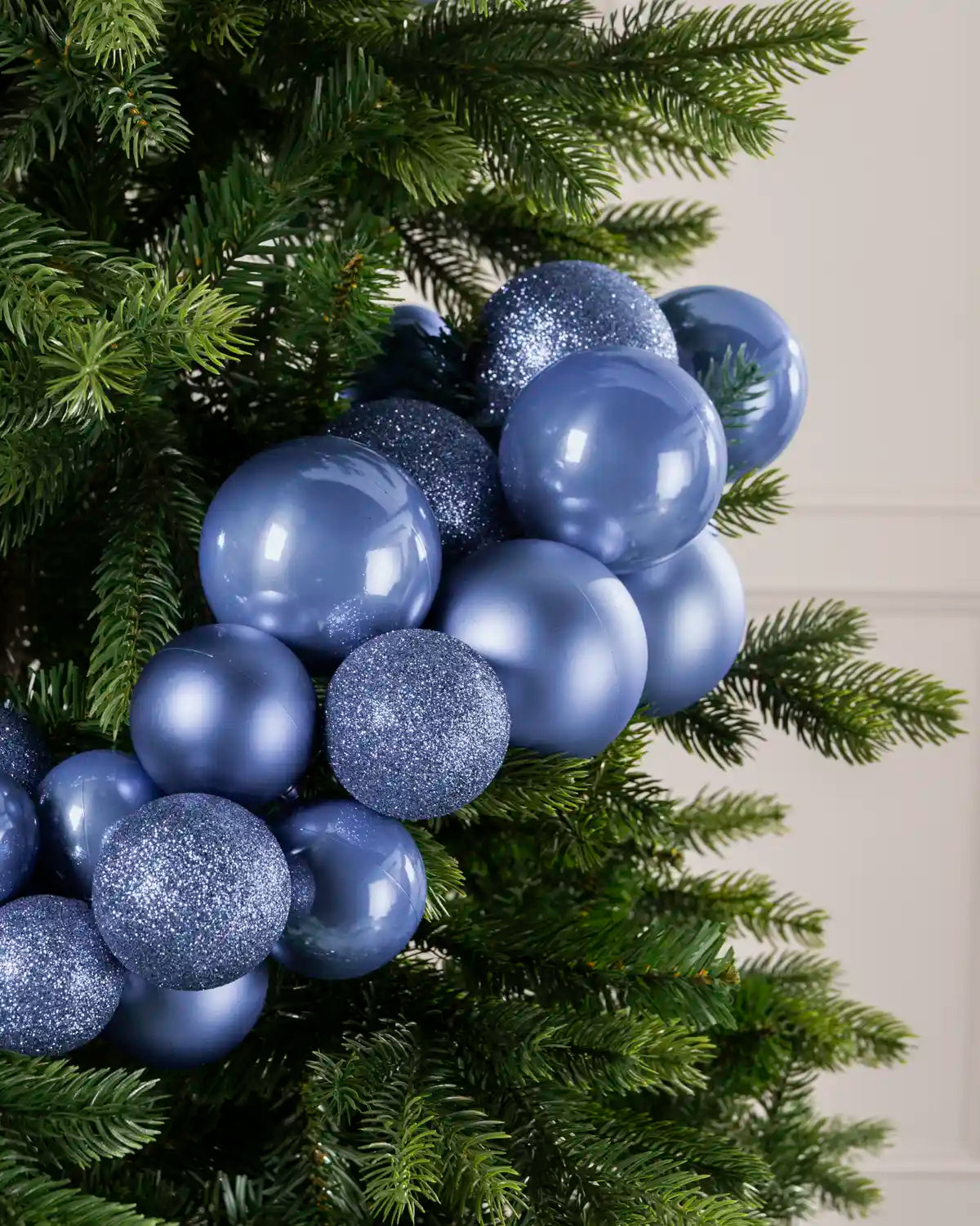 A cluster of Ice blue Christmas tree ornaments, including shiny matte blue balls, shiny glittery blue balls, and textured glitter blue balls, hanging on a green artificial Christmas tree.