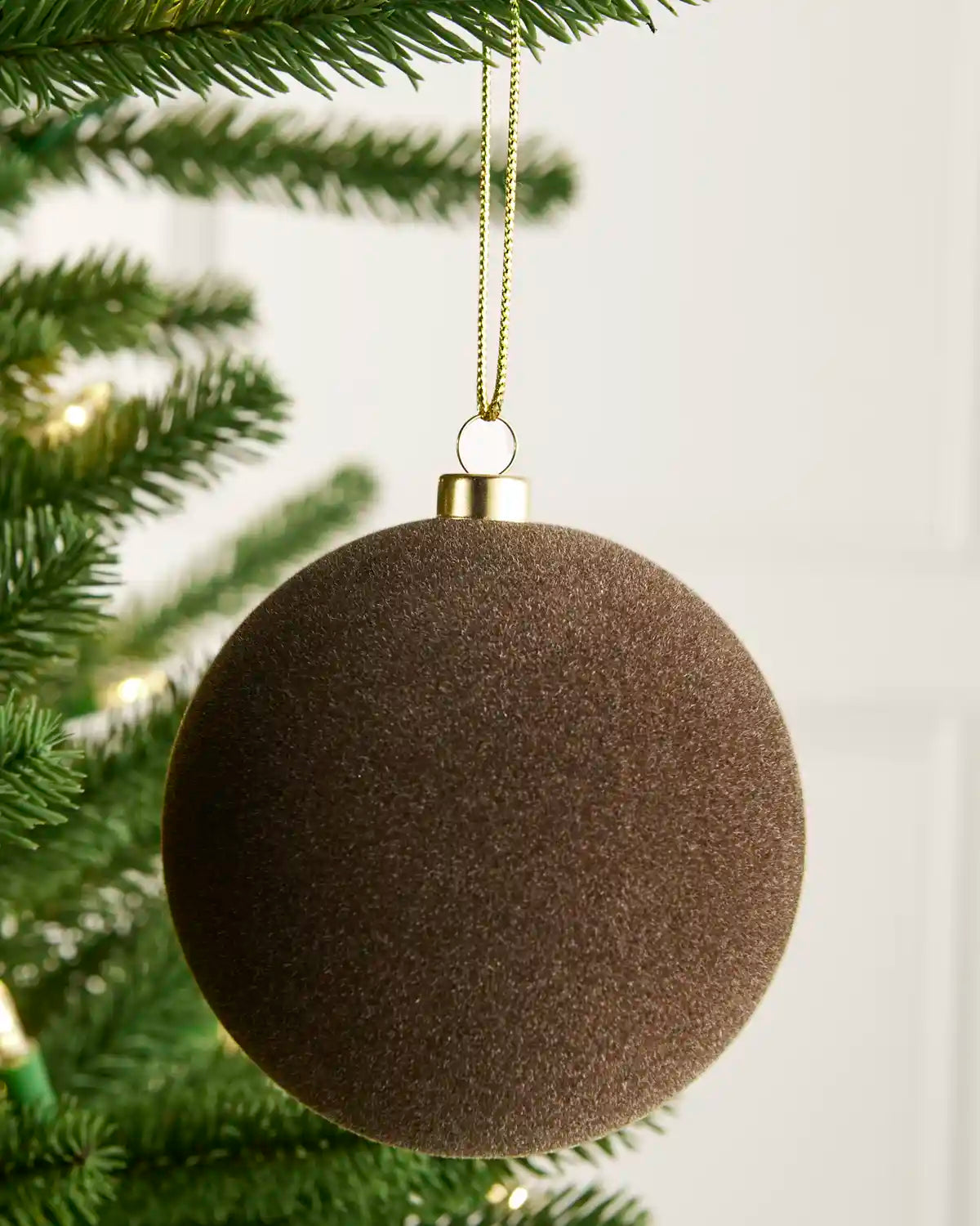 A dark brown textured spherical Christmas ornament hanging from a gold string on a green pine tree branch against a soft white background.
