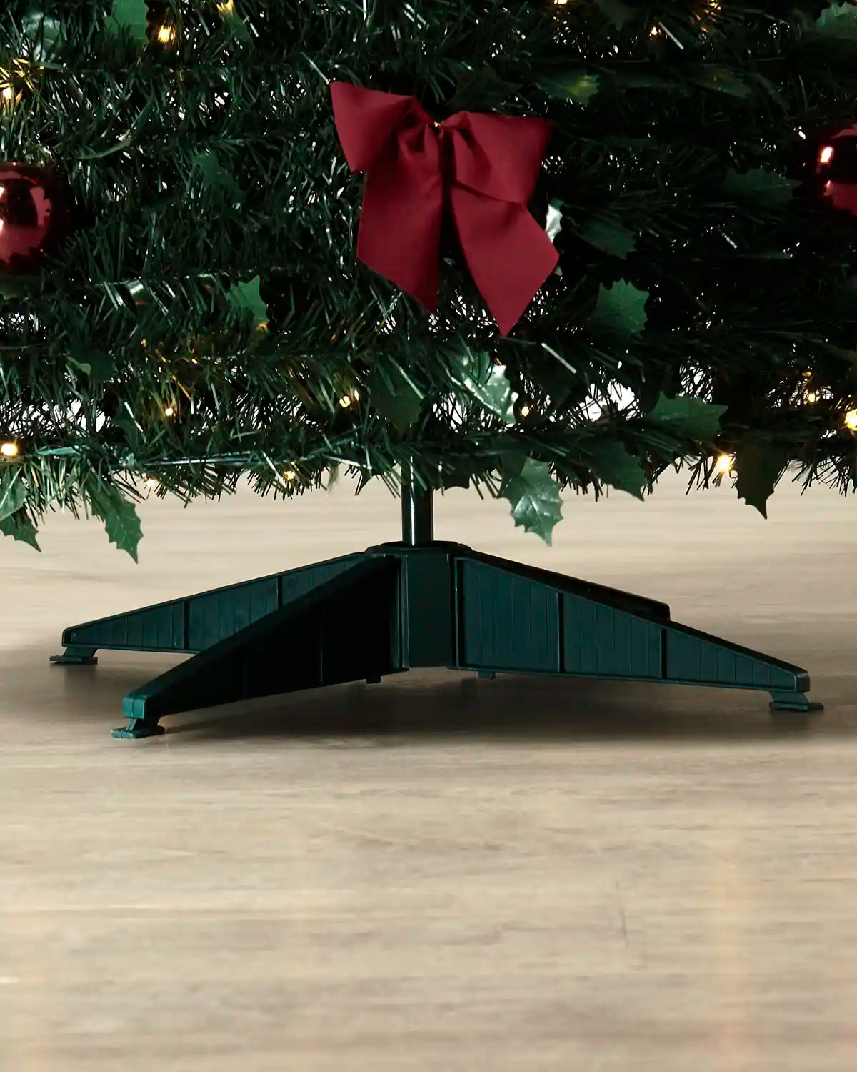 A dark green Christmas tree stand with a triangular base, shown supporting a decorated tree with a red bow and warm string lights on a light wood floor.