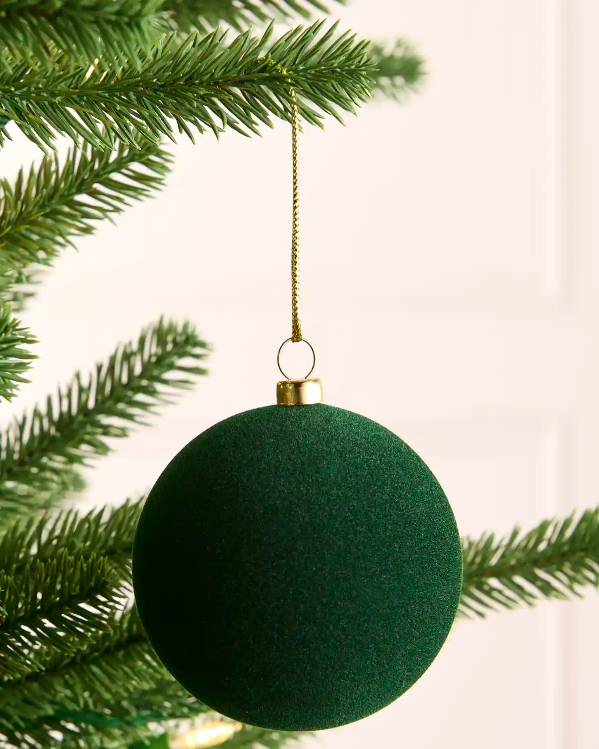 A dark green velvet Christmas tree ornament hanging from a gold string on a green pine tree branch against a soft off-white background.