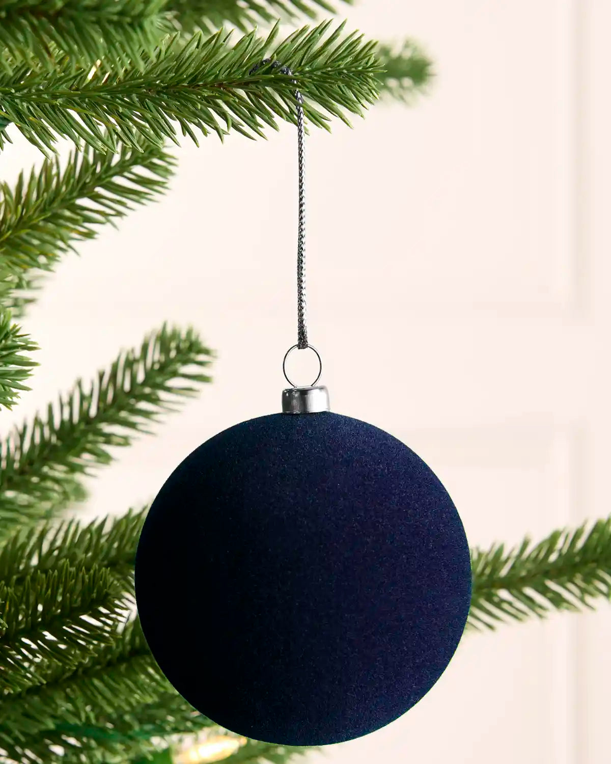 A dark navy blue spherical Christmas ornament hanging from a green pine tree branch against a soft off-white background.