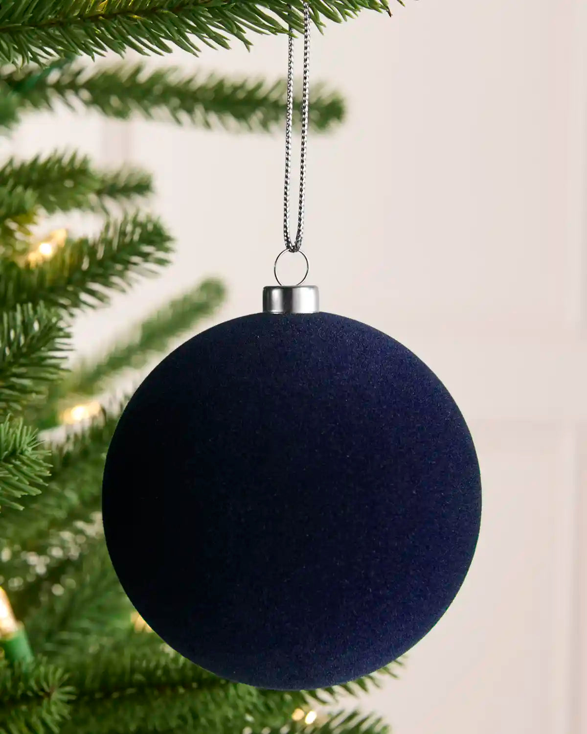 A dark navy blue spherical Christmas ornament hanging from a silver cap on a silver string, positioned in front of green pine tree branches with warm white string lights.
