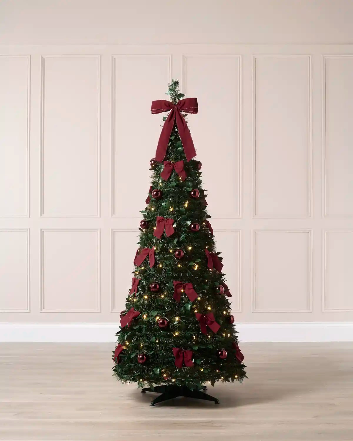 A decorated Holly Christmas tree with red ribbons, red ornaments, and warm white lights, standing on a black base against a cream panelled wall.