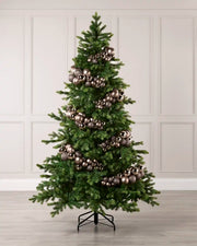 A decorated artificial Christmas tree with green needles and metallic mocha ornaments, standing on a black metal stand against a light gray paneled wall and wood floor.
